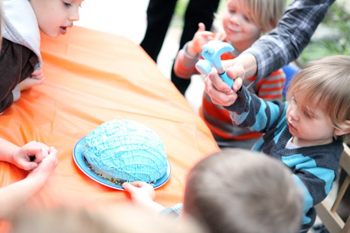 A round birthday cake on a table with kids around it. 