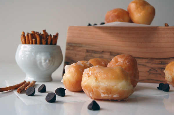 Ingredients to make chocolate and pretzel dipped donut holes on a table with a wooden block in the background. 