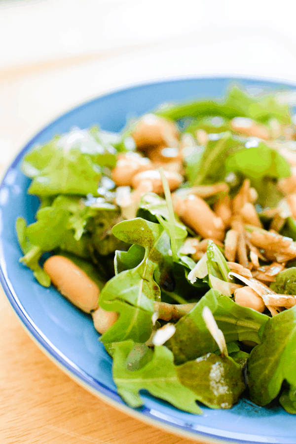 Close up of an arugula salad with white beans and parmesan in a blue bowl. 