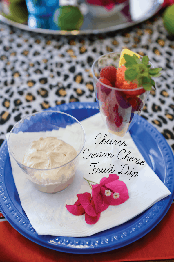 A cup holding Churro Fruit Dip on a plate with a flower and cup of fresh fruit to dip in it. 