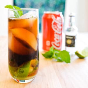 A cocktail on a table with a can of coke and small bottle of dark rum in the background.