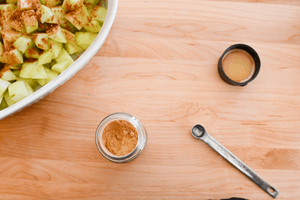 Cutting board with a bowl holding chopped green apples next to an open spice jar and measuring spoon. 