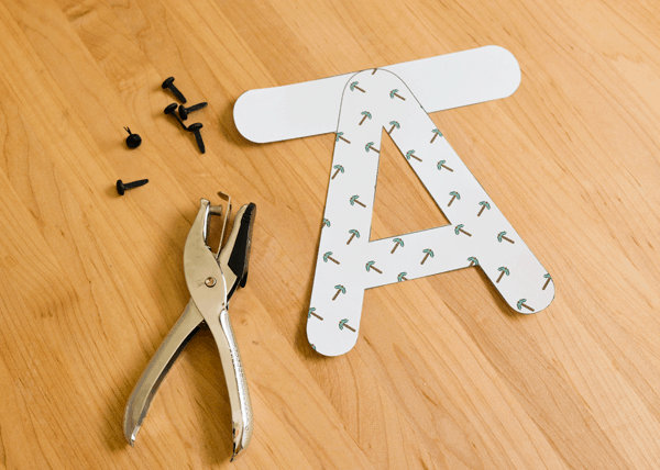 An "A" with tabs on the top cut out on a table next to brads and a hole puncher for a diy party banner. 