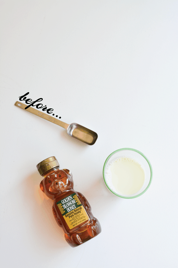 A bear shaped bottle of honey on a white table next to a measuring spoon and a glass of milk. 