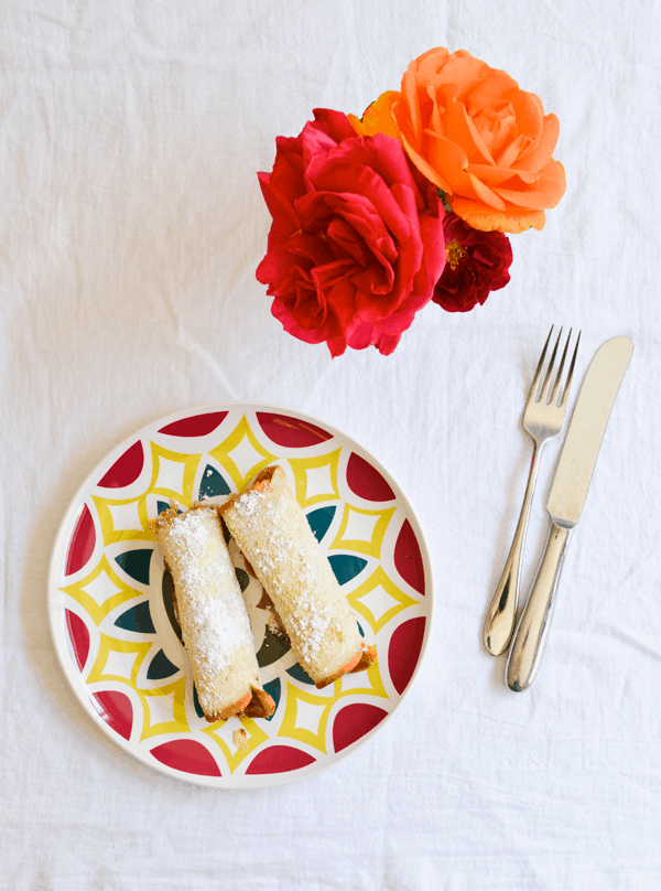 A white table with a plate holding two bread roll ups sprinkled with powdered sugar next to a knife and fork and a small vase of flowers. 