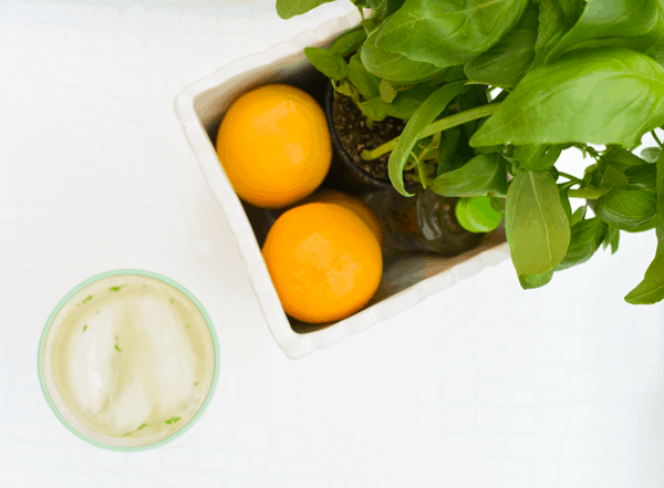 A small white planter holding a basil plant, two lemons and a small bottle of gin next to a cocktail on a table. 