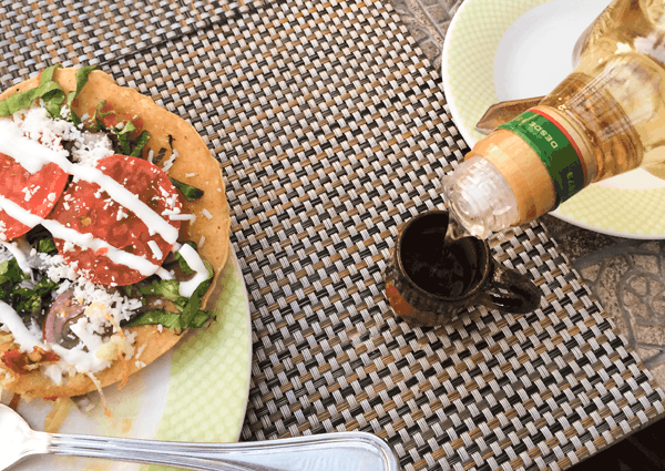 A tequila shot being poured with a tostada on a plate on the table next to it. 