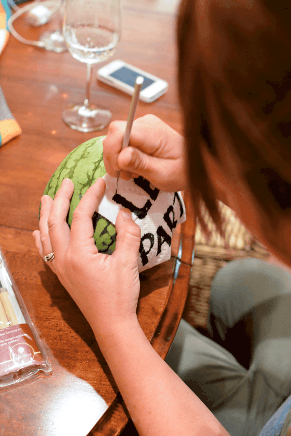 A woman using a tool to carve a sign on to a watermelon. 