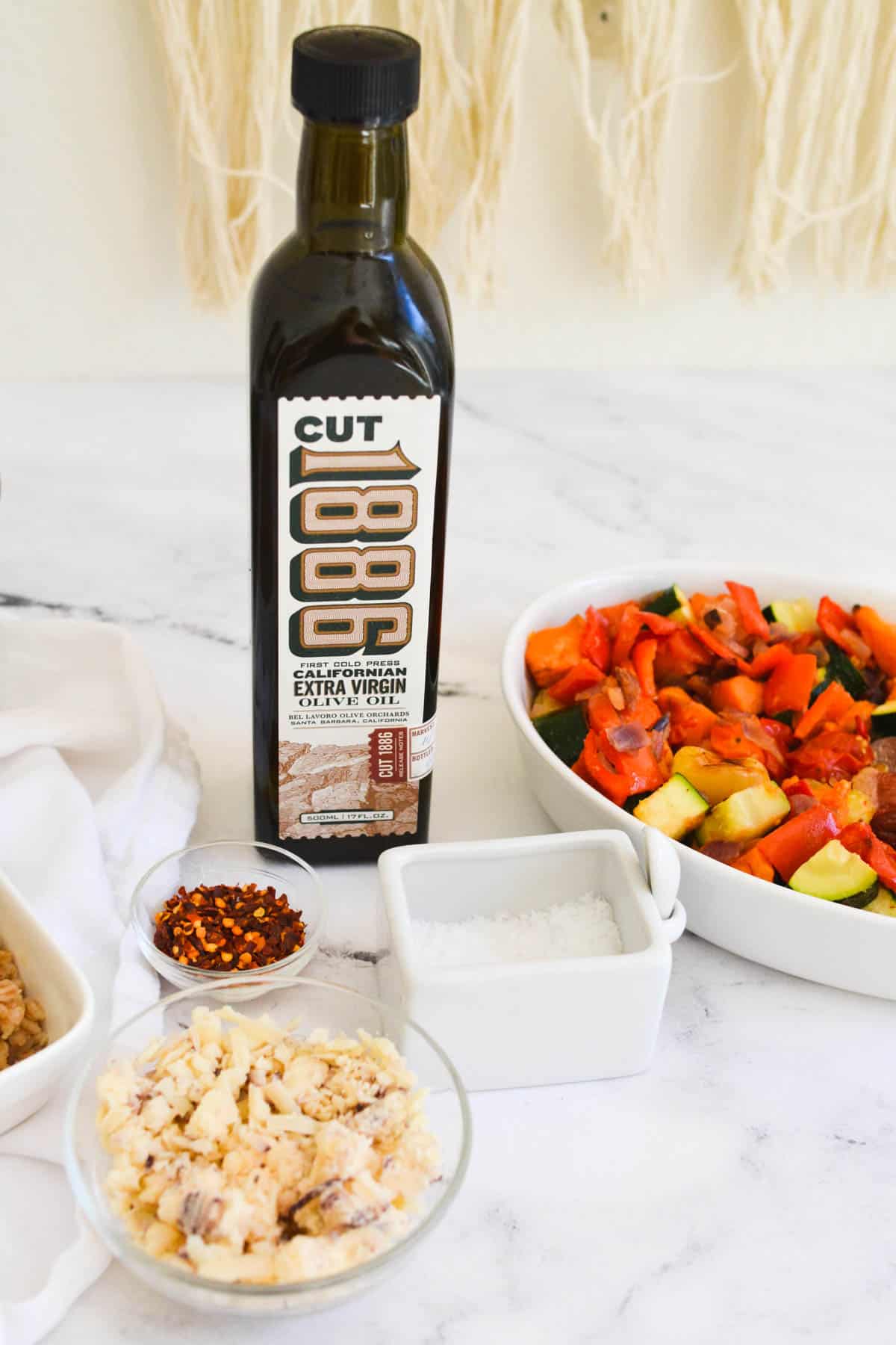 A bottle of olive oil on a table next to a bowl of roasted veggies, red pepper and salt.