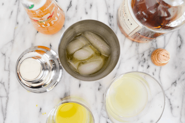 Overhead shot of a cocktail shaker with ice next to a bottle of bourbon, apple juice and a small dish of lemon juice.