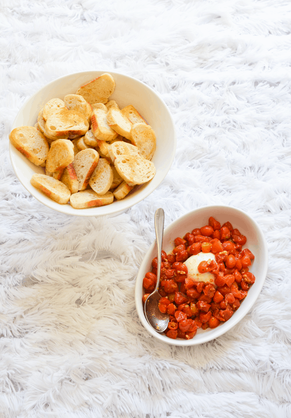 Roasted cherry tomatoes with garlic and burrata in a dish next to crostini for an easy party appetizer idea.