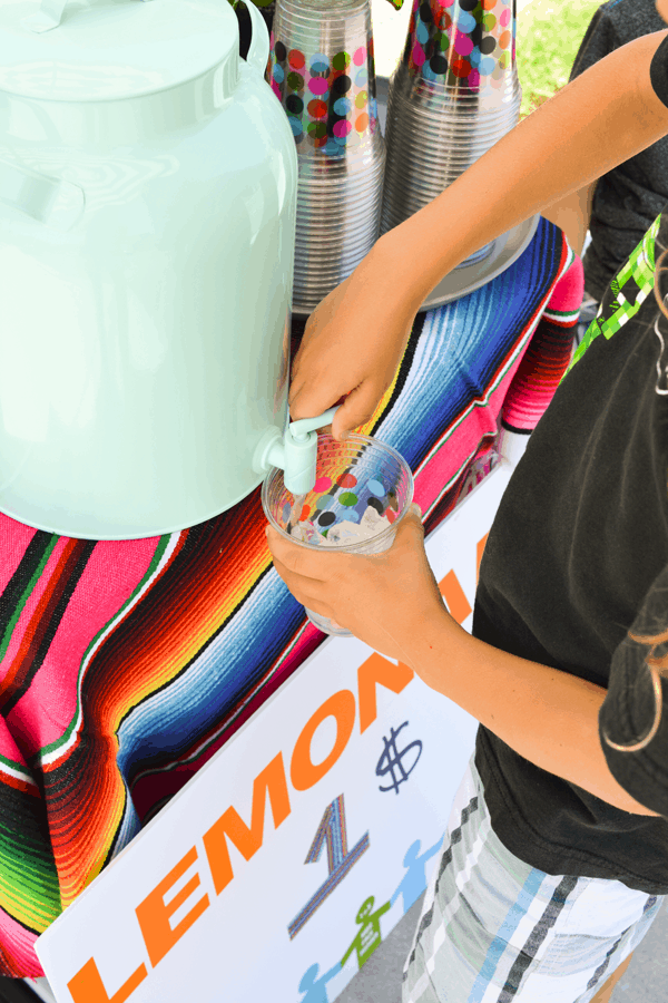 Kid adding lemonade to a cup of ice from a large batch of lemonade in a dispenser. 