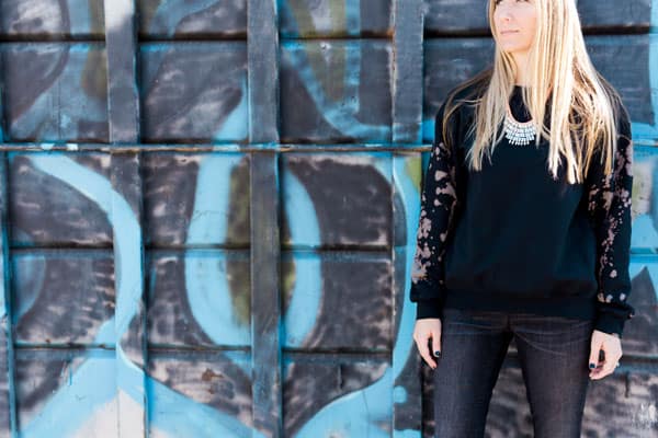 Girl wearing black sweatshirt with bleach splattered sleeves standing in front of graffiti.