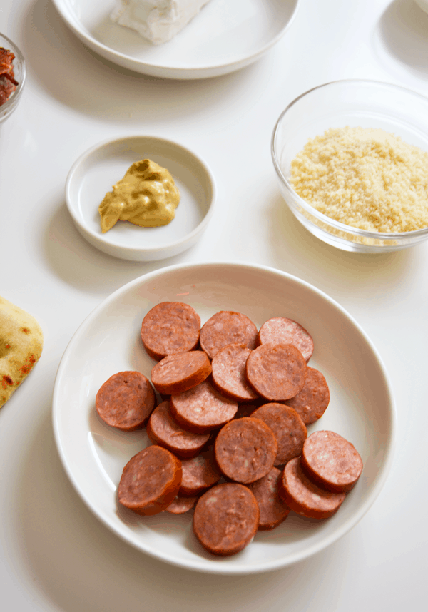 Sausage in a white dish next to a small plate filled with dijon mustard and a glass bowl with Parmesan cheese.