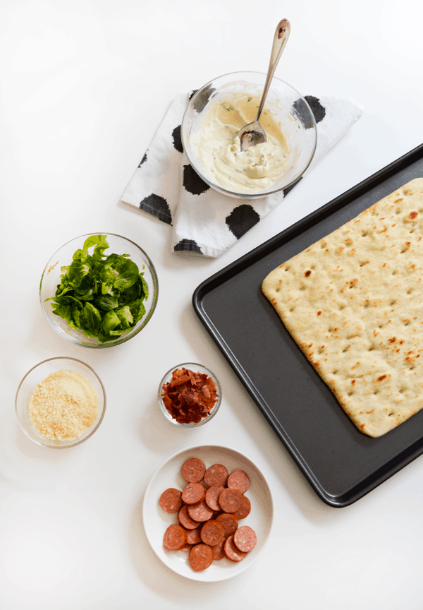 Ingredients in small dishes to make a Brussels sprouts and goat cheese pizza next to a baking sheet with flatbread pizza crust.