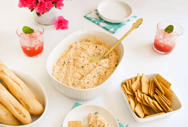 Dish with bacon cheese dip on a table next to bowls with crackers and baguettes and some drinks. 