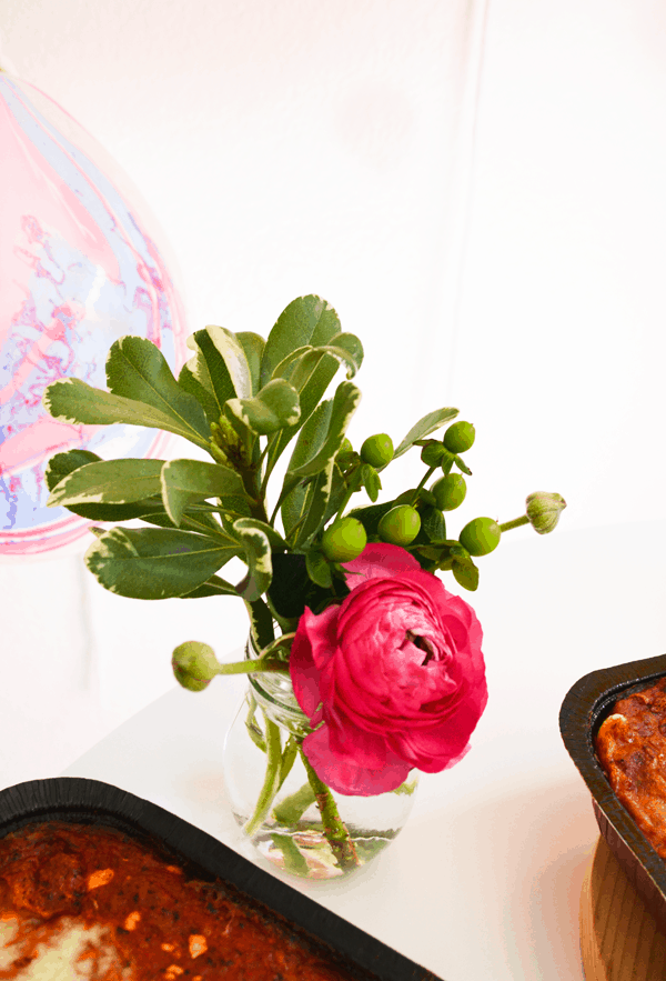 Pink fresh flower with leaves and green berries in a small glass vase on a party table by food.