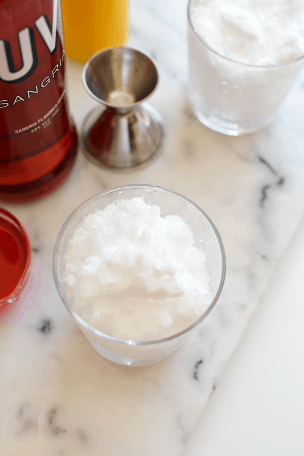 Glass of crushed ice on a counter next to a jigger and bottles of ingredients.