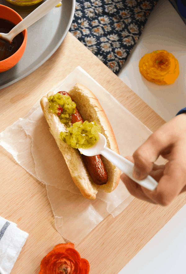 A kid putting relish on a hot dog with a white plastic spoon.