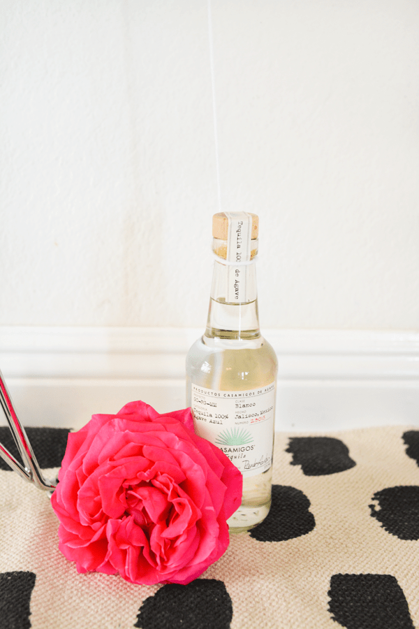 A bottle of tequila on the ground with strings of balloons tied to it and a large pink flower next to it. 