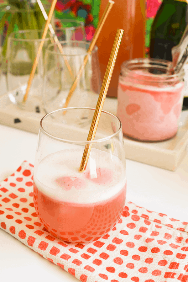 A stemless wine glass filled with a champagne float and a copper straw, on top of a red and white dotted napkin, in front of a tray holding ingredients.