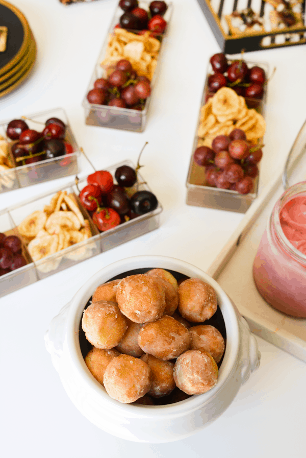 Glazed donut holes in a white turine in front of fresh and dried fruit in clear containers.