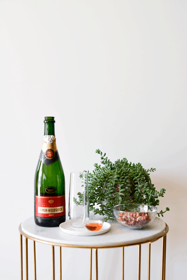 A bottle of champagne, a little bit of rose syrup, dried rose petals in a bowl and a champagne flute on a table. 