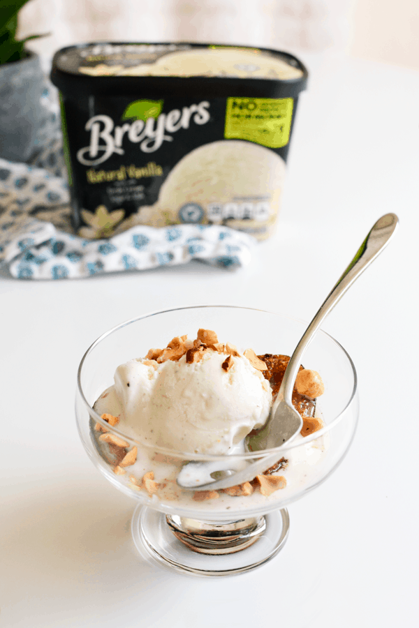 A glass bowl filled with roasted figs, vanilla ice cream, and toasted hazelnuts, with a spoon sitting in front of a carton of vanilla ice cream.