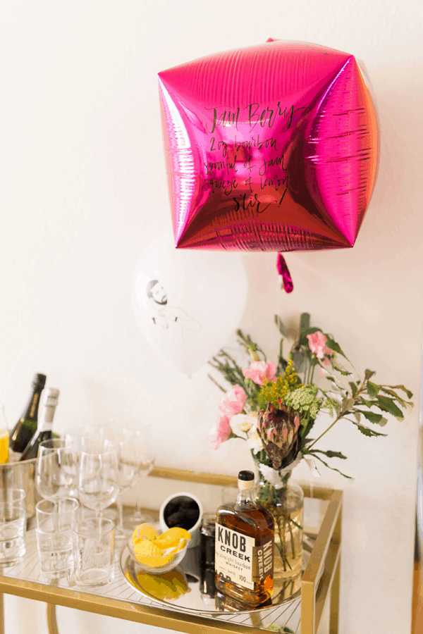 A cocktail recipe written in cursive on a mylar balloon attached to a bar cart with ingredients on it.