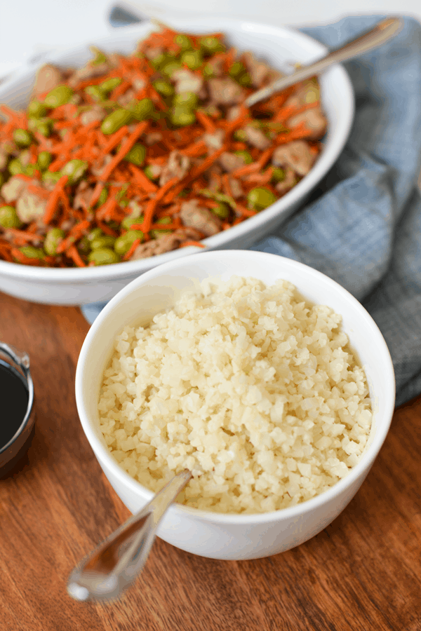 White bowl of riced cauliflower on a wood tray next to a bowl of ground turkey teriyaki stir fry for dinner.