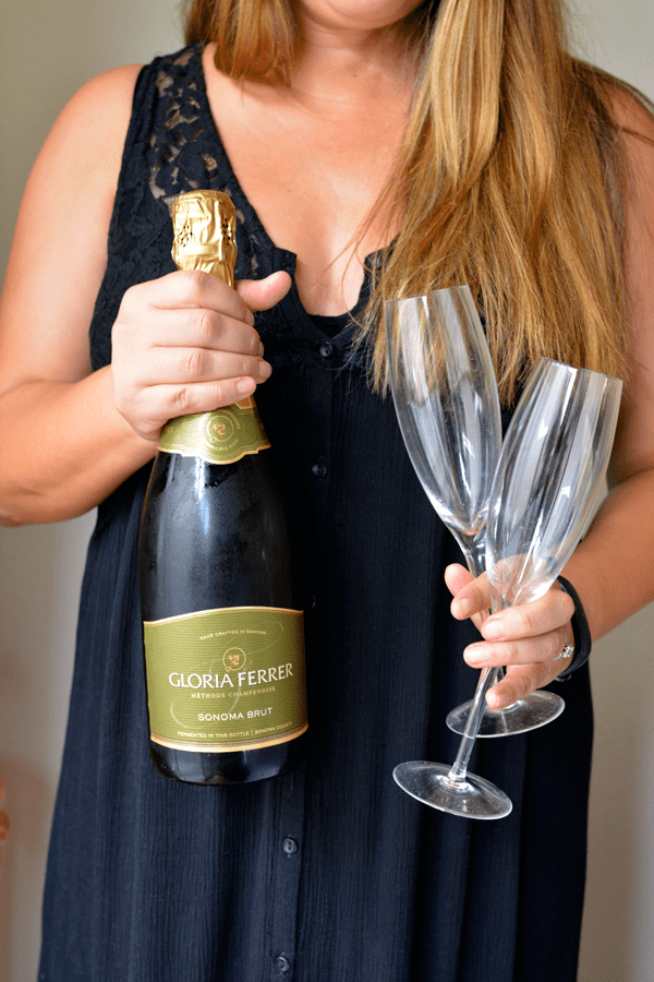 Woman holding a bottle of Gloria Ferrer champagne in one hand and two glass champagne flutes in the other hand.