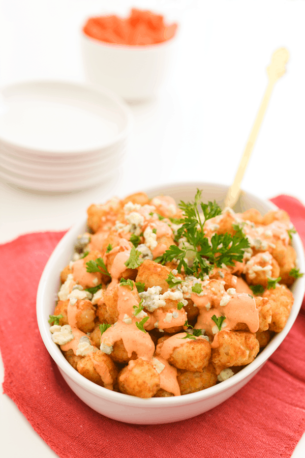 A white baking dish holding loaded Tater Tots with Buffalo Aioli and blue cheese crumbles next to appetizer plates. 