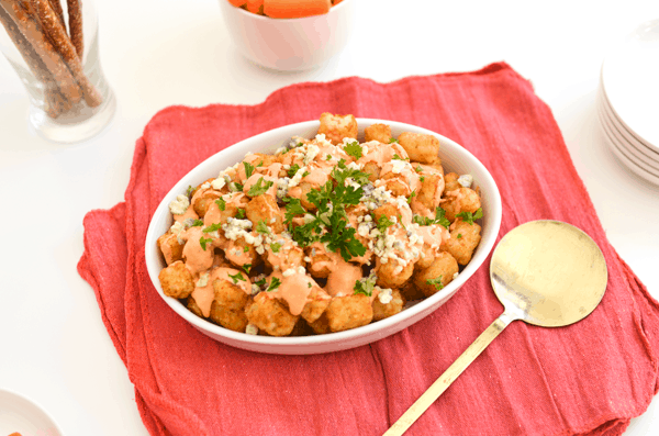 Loaded tater tots topped with blue cheese crumbles, parsley and Buffalo Aioli in a white serving dish. 