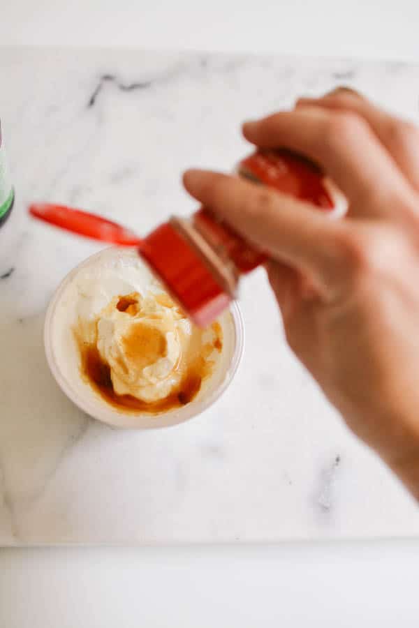 A hand sprinkling cinnamon from the jar into a tub of Greek yogurt.