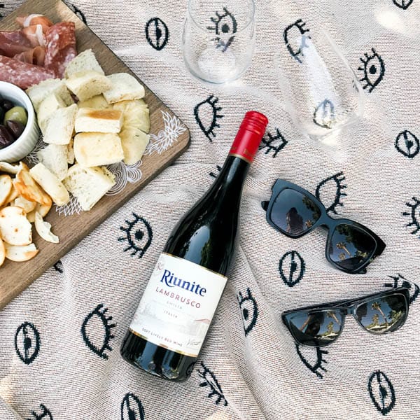 A bottle of Riunite Lambrusco laying on a blanket next to a charcuterie board and mens and womens sunglasses.