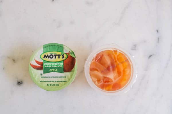 A cup of applesauce and a small fruit cup on a table. 