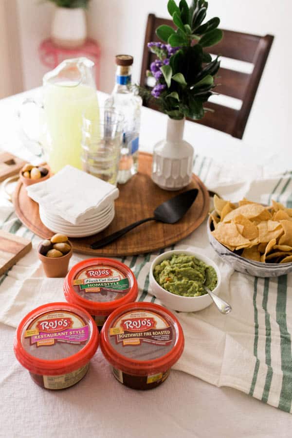 A dining table with party food and 3 containers of salsa next to guacamole and a bowl of chips. 