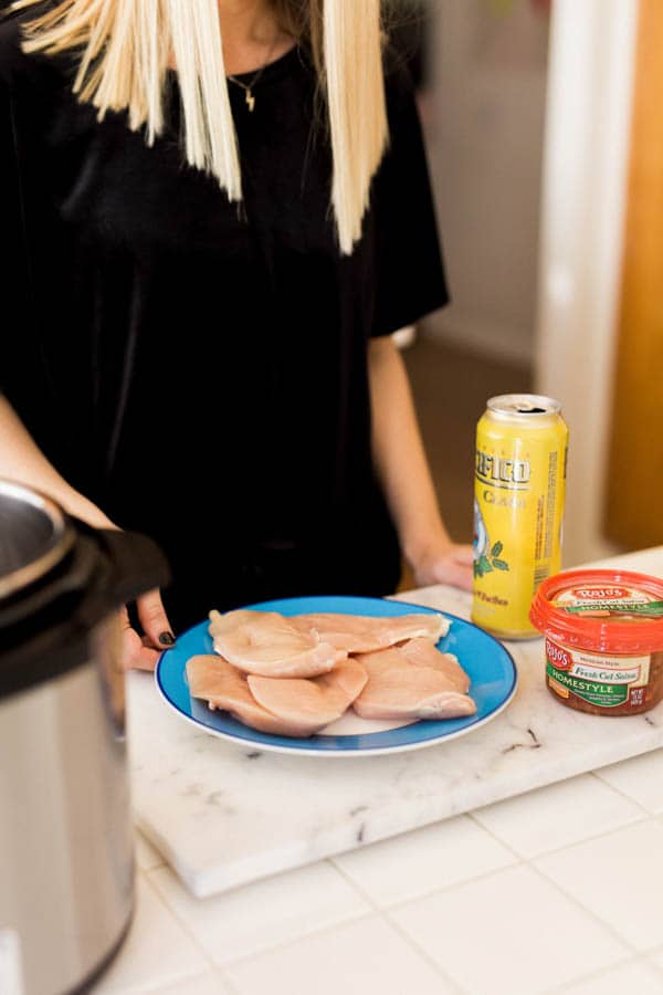 Woman standing in front of a can of beer, pot of salsa and sliced chicken breasts on the kitchen counter.