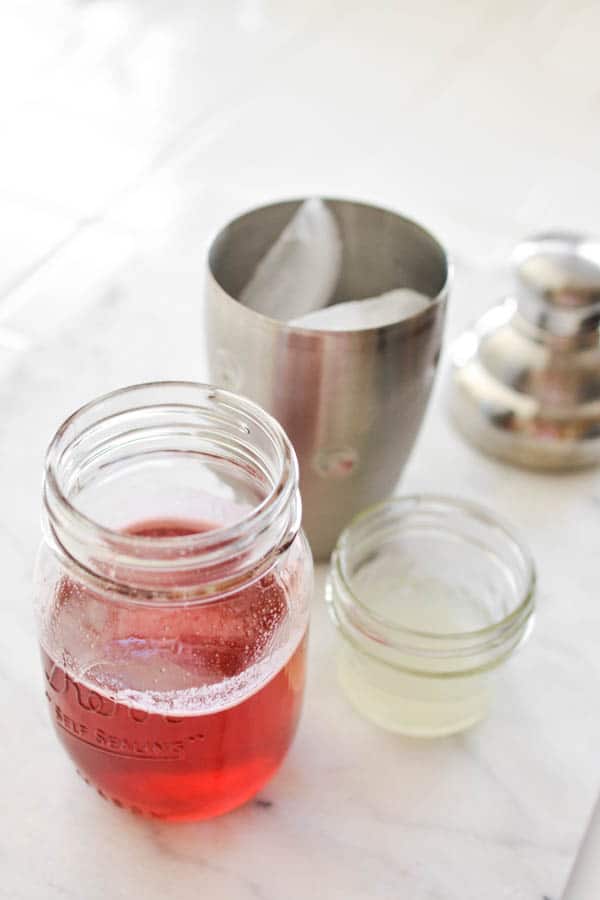 Strawberry simple syrup and lime juice next to a cocktail shaker on a table.