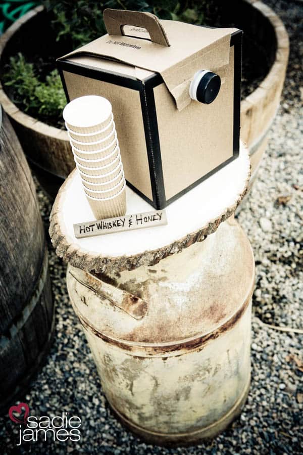 A chipboard insulated drink container and hot drink cups on a wooden table with a wooden sign that says Hot Whiskey and Honey.