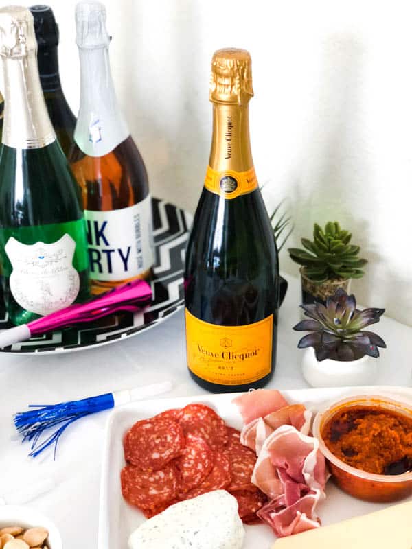 A bottle of champagne on a party table next to a charcuterie plate and noise blowers for New Year's Eve.