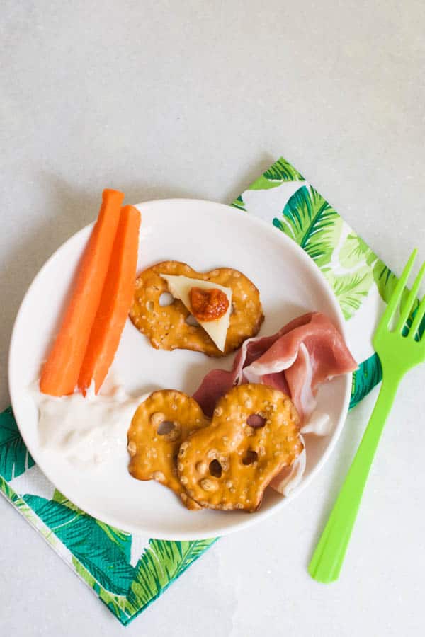 Pretzel crisps on a small appetizer plate with dip, cheese, carrots and meat next to a napkin and plastic fork. 