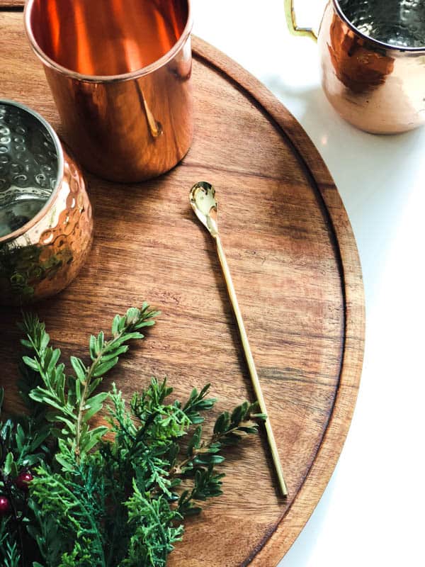 A wooden tray topped with copper mugs, a cocktail spoon, and greenery. 