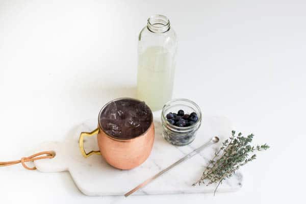 Ingredients for a Blueberry Lemonade Moscow Mule with the finished cocktail on a white marble board and a neutral background.