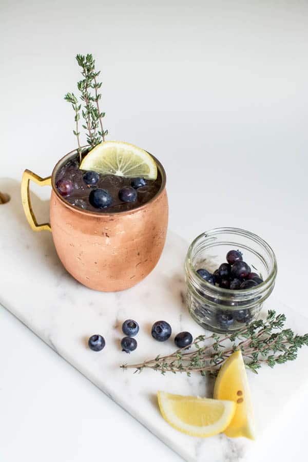 Blueberry lemonade Moscow Mule cocktail in a copper mule mug pictured among spare ingredients on a marble board with a neutral background.