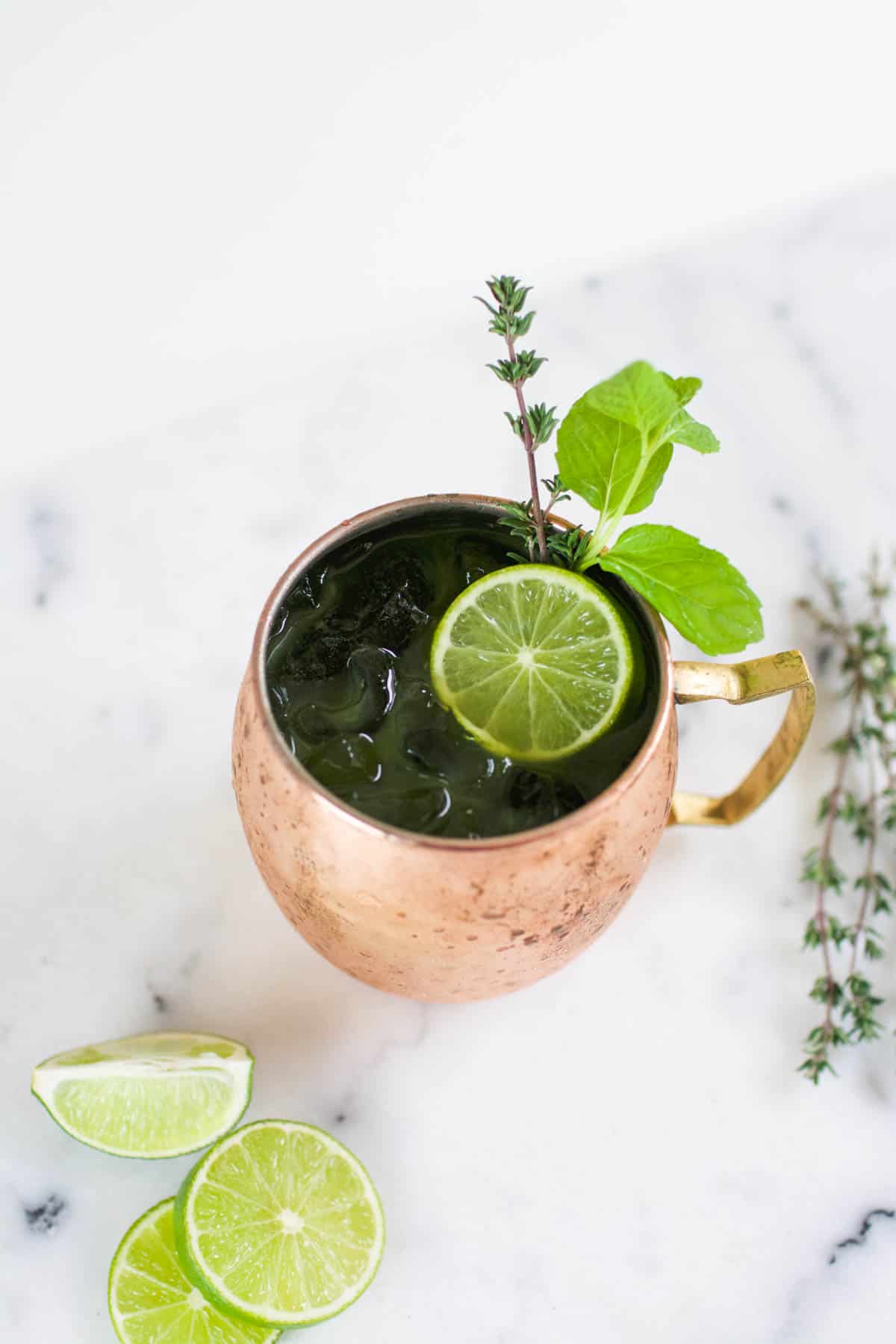 Copper Mule mug on a table holding a cocktail with limes and fresh herbs in it and around it.