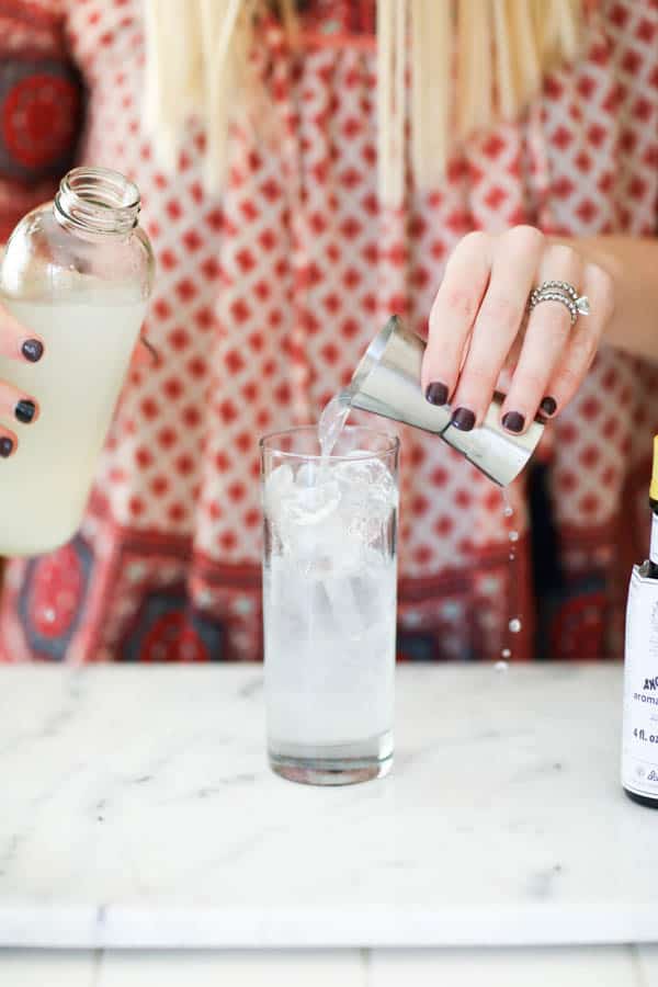 Woman pouring lemonade from a jigger into a tall cocktail glass with ice. 