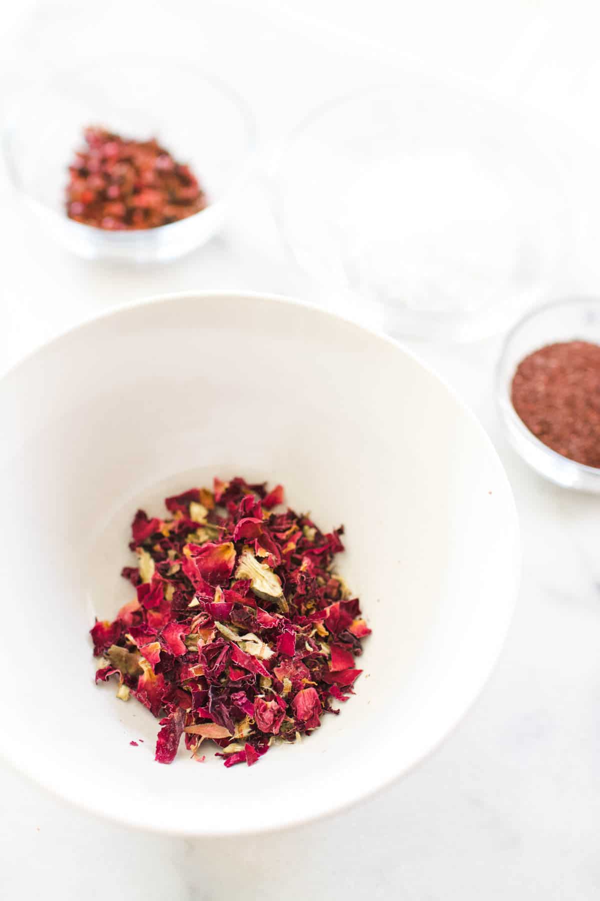 A bowl with dried rose petals on a counter next to small bowls with other spices.