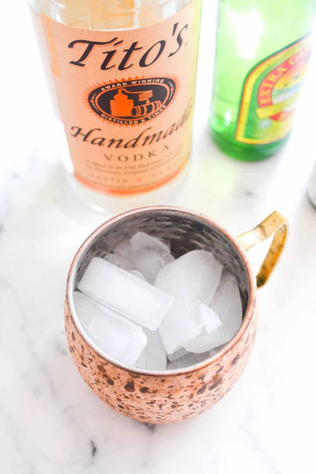 Close up of a copper mug holding ice on a counter next to a bottle of vodka and ginger beer.