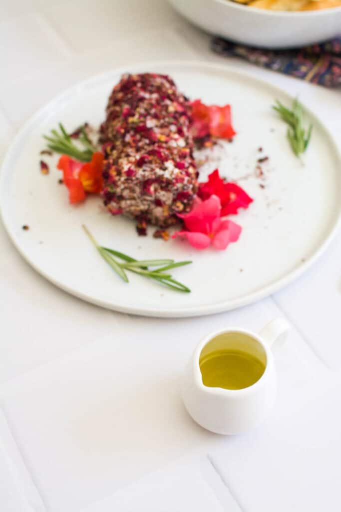 A spice crusted goat cheese log appetizer on a plate garnished with edible flowers with a small pitcher of olive oil next to it on a table.